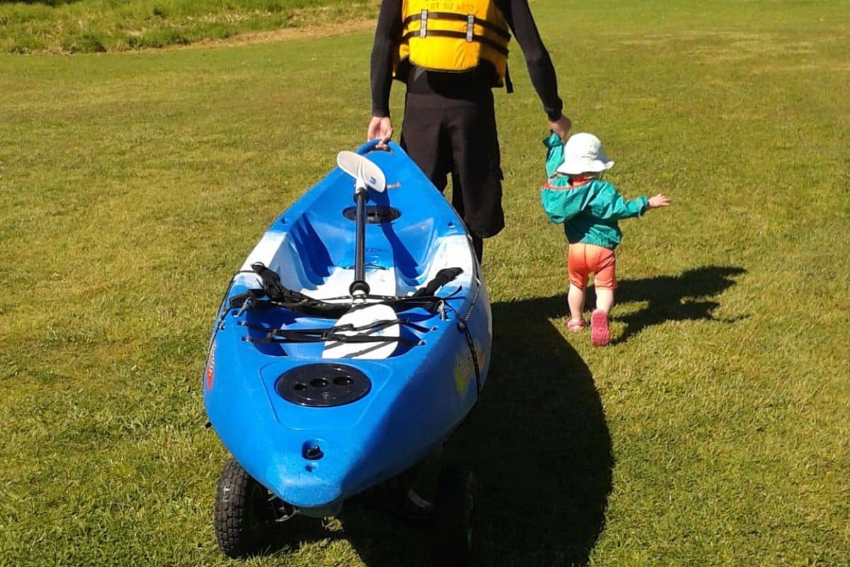 Man pulling a blue kayak on a cart while holding a child
