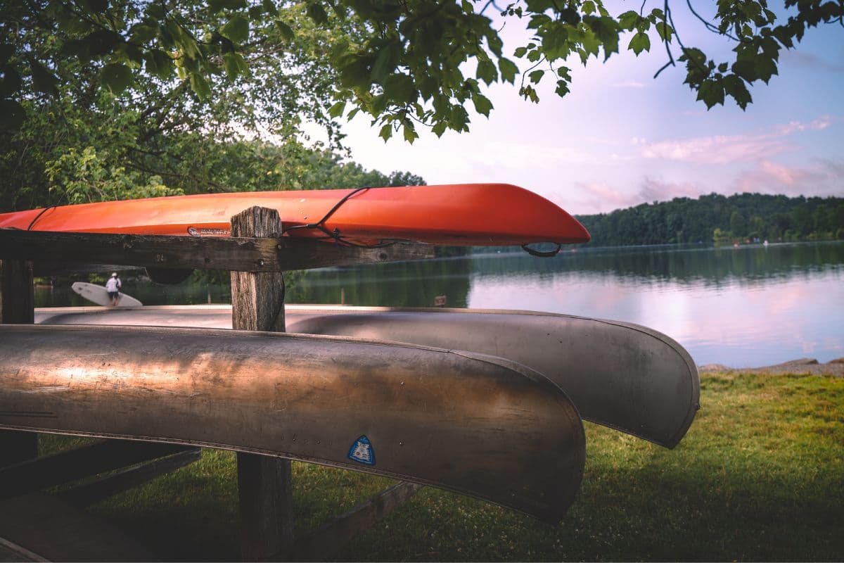 Two wooden kayaks and a red ABS kayak displayed on a rack by the lakeside